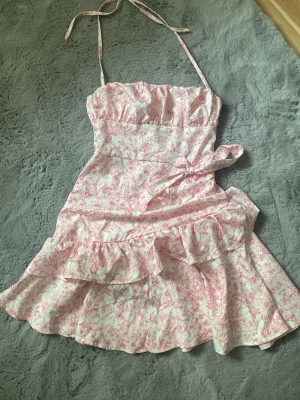 Rosa blommig kortklänning med volang - kortklänning med rosa blommönster på vit botten. Klänningen har tunna axelband, volangdetaljer och ett knyt i midjan för extra form.