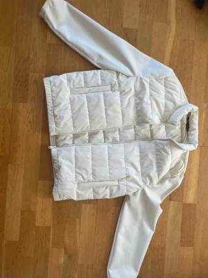 Vit cardigan från Zara - Snygg vit pufferjacka från Zara med raka linjer och quiltad design. Jackan har hög krage, dragkedja framtill och två praktiska fickor med dragkedja. Tillverkad i ett glansigt material som ger en modern look. Perfekt för kalla vinterdagar. Har använt bara en gång. Därför jag säljer den