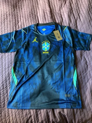 Nike Brasil blå matchtröja - Snygg blå och mörkgrön matchtröja från Nike med Brasil-logga och gula detaljer. Tröjan har korta ärmar, rund hals och är tillverkad i ett lätt, ventilerande funktionsmaterial. Perfekt för fotboll eller träning.