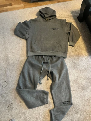 Grått loungewear-set Essentials - *Pris diskuteras privat*Säljer ett par grå mjukisbyxor från Essentials Fear of God med svart logga på benet. Byxorna har dragsko i midjan och är tillverkade i mjukt bomullsmaterial för skön känsla. Size L. Perfekta för en avslappnad och trendig look. Även med en matchande Hoodie med Size M.