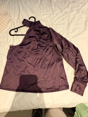 Vinröd oneshoulder blus med hög krage - Snygg vinröd oneshoulder blus i glansigt satintyg. Blusen har en lång ärm med manschett och knappar samt en hög krage med dekorativa knappar. Den är aldrig använd endast testad!!
