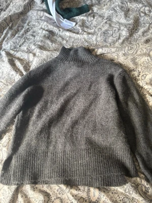 Grå stickad polotröja - Mysig grå stickad polotröja med bred ribbad mudd vid hals, ärmslut och nederkant. Tröjan har en avslappnad passform och är perfekt för kyliga dagar. Materialet ser ut att vara ull eller en ullmix för extra värme.