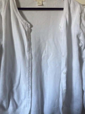 Vit basic kofta från H&M - Klassisk vit kofta från H&M med rund halsringning och knäppning framtill. Tillverkad i mjukt, tunt stickat material med ribbade muddar vid ärmslut och nederkant. Perfekt att slänga över en topp eller klänning för en clean look.