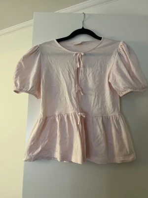 Ljusrosa blus med puffärm och knyt - Ljusrosa blus med puffiga kortärmar och tre dekorativa knyt framtill. Blusen har volang nedtill och en lös, avslappnad passform som ger en söt och romantisk look. Perfekt för dig som gillar mjuka färger och feminina detaljer.