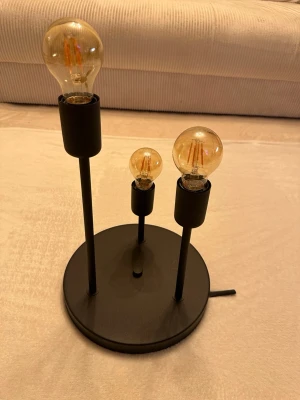 Svart taklampa med tre glödlampor - Modern taklampa i svart metall med rund fot och tre olika höga lampfästen. Lampan har tre synliga glödlampor med varm ton som ger en industriell och stilren känsla. Perfekt som inredningsdetalj i vardagsrum eller sovrum. Ljuskällor ingår. Oanvända!