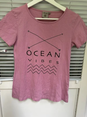 Rosa t-shirt med tryck från Tribute storlek: M - Snygg rosa t-shirt från Tribute med svart tryck som visar två korsade pilar, texten 'OCEAN VIBES' och vågmönster. T-shirten har rund halsringning och korta ärmar. Perfekt för en avslappnad stil.