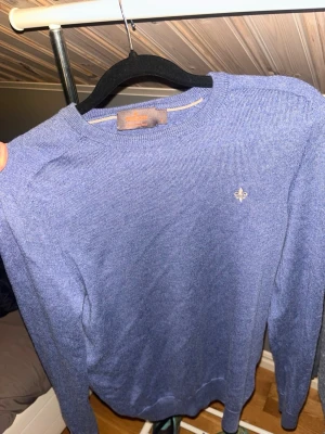 Blå stickad tröja från Morris - Snygg blå stickad tröja från Morris med rund halsringning och diskret logga broderad på bröstet. Tröjan har långa ärmar och är tillverkad i ett tunt, mjukt material som passar perfekt till lager på lager. Enkel och stilren design.