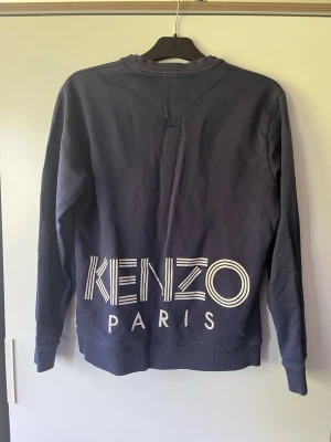 Mörkblå sweatshirt från Kenzo Paris - Snygg mörkblå sweatshirt från Kenzo Paris med stort vitt tryck på ryggen. Tröjan har rund halsringning, ribbade muddar och är tillverkad i mjukt bomullsmaterial. Perfekt för en avslappnad och trendig stil.
