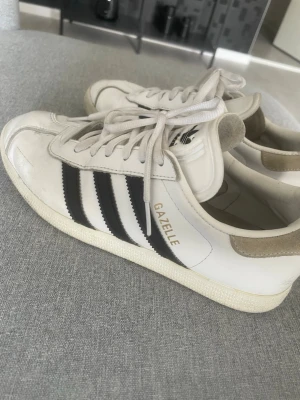 Adidas Gazelle vita sneakers - Klassiska Adidas Gazelle sneakers i vitt läder med svarta ränder på sidorna och guldfärgad 'GAZELLE'-text. Skorna har låga skaft, vita snören och beige mockadetalj vid hälen. Perfekta för en sportig och stilren look. Limmet har släppt lite men går att limma därav priset.