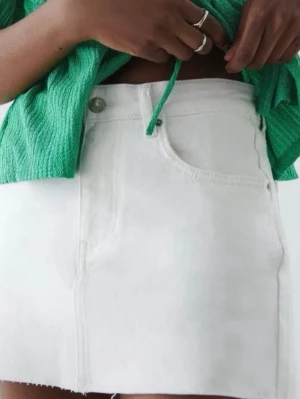 Vit jeanskjol med rå kant - Säljer en vit kort jeanskjol med rå, fransig kant nertill och klassiska femficksdetaljer. Kjolen har knapp och dragkedja framtill och är perfekt för en trendig och avslappnad look.