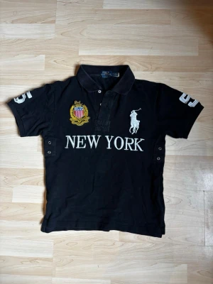 Svart piké från Polo Ralph Lauren - Svart pikétröja från Polo Ralph Lauren med broderad logga och texten NEW YORK på bröstet. Har vita detaljer, broderat emblem och knappar vid halsen. Klassisk krage och korta ärmar med siffror på båda ärmarna. Är storlek M men den sitter som en S 