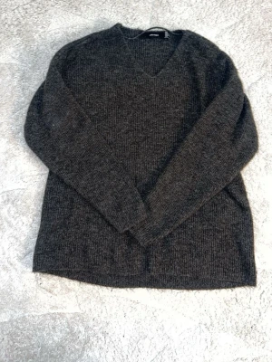 Mörkgrå stickad tröja Vero Moda - Mysig mörkgrå stickad tröja från Vero Moda med v-ringning och långa ärmar. Tröjan har en klassisk rak passform och är tillverkad i ett mjukt, ribbstickat material som passar perfekt för kyliga dagar.