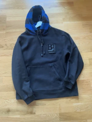 Svart Burberry hoodie med blå huva - Snygg svart hoodie från Burberry med stor ficka framtill och en huva i blått och svart rutmönster. Bröstet har en upphöjd B-detalj. Dragsko i huvan och ribbade muddar vid ärmar och nederkant. Perfekt för dig som vill ha något stilrent men ändå unikt. Inga defekter förutom det lilla hålet, kan sys ihop enkelt. Köpt för 900 dollar så riktigt prisvärt!