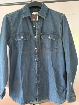 Blå jeansskjorta från Levi's - Klassisk blå jeansskjorta från Levi's med långa ärmar, två bröstfickor med knappar och vit knappstängning framtill. Skjortan har en avslappnad passform och är gjord i slitstarkt bomullsmaterial. Perfekt till jeans eller kjol för en cool denimlook. Ordinariepris 900kr, säljer för 500kr.