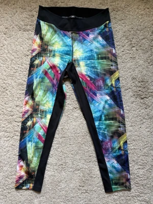 Färgglada träningsleggings från SOC - Helt nya och oanvända. Möjligen tvättade en gång. Säljer ett par leggings från SOC med färgstarkt grafiskt mönster i blått, grönt, rosa och gult. Svart midjeresår med SOCX89-tryck bak och svarta paneler längs benen. Stretchigt material som sitter skönt och följsamt.