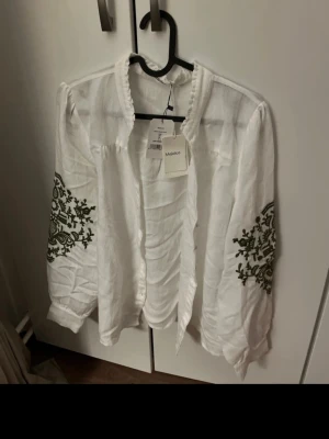 Vit blus med broderi från Majolica - Säljer en luftig vit blus från Majolica med grönt broderi på ärmarna. Blusen har lång ärm, volangkant vid kragen och är gjord i ett tunt, skönt material. Perfekt för dig som gillar boho-vibbar och detaljerade mönster.
