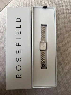Silverfärgad klocka från Rosefield - Stilren klocka från Rosefield med fyrkantig urtavla och länkarmband i silverfärg. Urtavlan är vit med minimalistisk design och tunna visare. Perfekt accessoar för dig som gillar en klassisk och elegant look. Aldrig använd!!