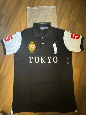 Polo Ralph Lauren - Vintage Chief keef polo - Svart pikétröja från Polo Ralph Lauren med vita ärmar, stora broderade detaljer och texten TOKYO på bröstet. Klassisk krage, knappar framtill och röda siffror på ärmarna. Perfekt för dig som gillar sportig och stilren look.