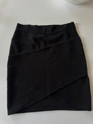 Svart omlottkjol i ribbad stil - Säljer en svart kortkjol med omlottdetalj framtill och ribbad struktur. Kjolen har en enkel, stilren design och passar perfekt till många olika looks. Materialet känns mjukt och stretchigt för en bekväm passform.