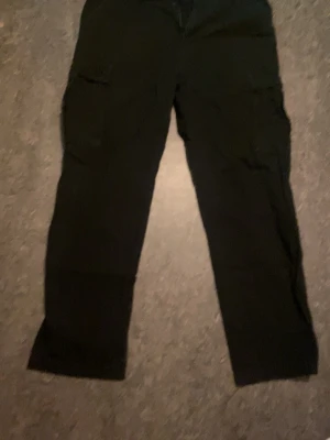 Svarta cargobyxor med fickor - Säljer ett par svarta cargobyxor med raka ben och klassiska sidofickor på låren. Byxorna har en avslappnad passform och är perfekta för en streetwear-look. Materialet ser ut att vara bomull eller en bomullsblandning. Inga synliga loggor eller märken.