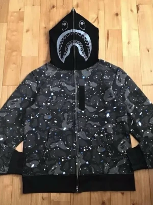 BAPE Shark Camo zip hoodie svart/grå - Säljer en BAPE Shark zip hoodie i svart och grått camomönster med vita och blå detaljer. Huvan har den ikoniska hajdesignen med tänder och ögon. Dragkedja hela vägen upp och två fickor fram. Ribbade muddar och nederkant. Perfekt för dig som gillar streetwear.