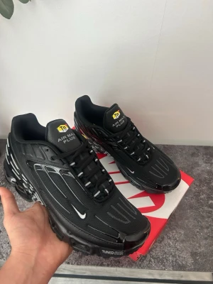 Nike Air Max Plus svarta sneakers - Nike Air Max Plus sneakers i helsvart med vita detaljer och TN-logga på plösen. Skorna har en sportig design med synliga luftkuddar i sulan och tunna linjer längs ovandelen. Materialet är en mix av mesh och syntet för både stil och komfort. Perfekta för dig som gillar streetwear.