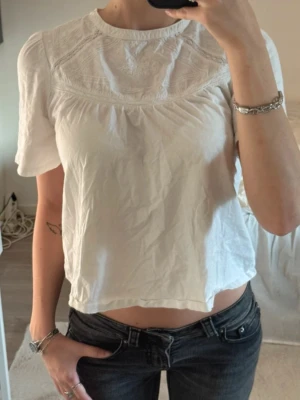 Vit blus med broderad detalj - Söt vit blus med korta ärmar och rund halsringning. Blusen har fina broderade detaljer upptill och ett lätt, luftigt tyg som ger en avslappnad känsla. Perfekt till jeans eller kjol för en enkel men stilren look. Passar som en S