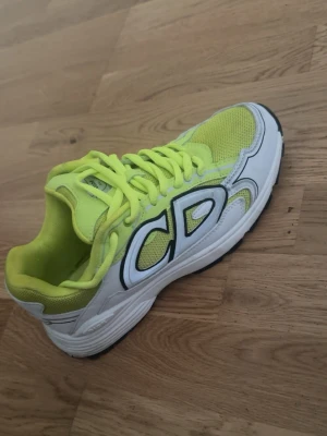 Gula och vita CD sneakers - Snygga sneakers i gult och vitt med stora CD-bokstäver på sidan. Ovandelen är i mesh och syntetmaterial, vilket ger bra andningsförmåga. Skon har en chunky sula och gula skosnören som matchar designen. Perfekt för dig som gillar streetwear och vill sticka ut.