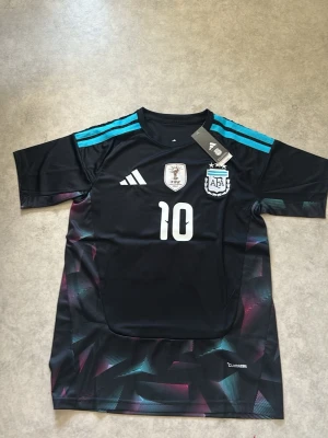 Argentina bortatröja Adidas nr 10 - Snygg svart bortatröja från Adidas för Argentinas landslag med Messi och nummer 10 på bröstet. Tröjan har turkosa detaljer på axlarna, broderade märken och CLIMACOOL-material som andas. Perfekt för fotbollsträning eller att bära till vardags.