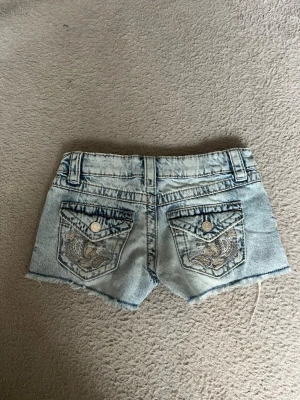 Ljusblå jeansshorts med broderi - Säljer ett par ljusblå jeansshorts med snygga broderade detaljer på bakfickorna och råa kanter. Shortsen har normal passform och bälteshällor. Perfekta för sommaren! Använda ca 3 gånger🩷🩷