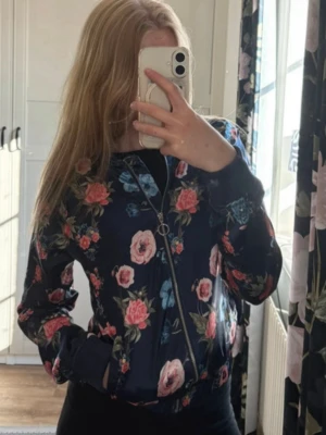 Blommig bomberjacka från H&M - Mörkblå bomberjacka från H&M med färgglatt blommönster i rosa, blått och grönt. Jackan har dragkedja framtill, ribbade muddar och sidofickor. Perfekt för dig som vill sticka ut med en snygg och trendig look.