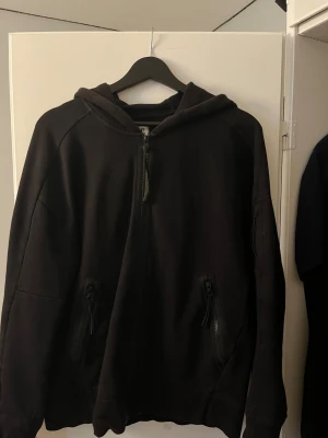 Svart hoodie från C.P. Company - Säljer en svart hoodie från C.P. Company med dragkedja framtill och två fickor med dragkedja. Använt skick, solbränd annars fin och hel! Tveka inte att höra av dig vid frågor!