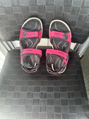 Rosa sportiga sandaler från Ecco - Snygga och bekväma sandaler från Ecco i svart och rosa med justerbara kardborreband. Sulan är robust och har bra grepp, perfekt för äventyr och vardag. Tillverkade i slitstarkt material med sportig design och kontrastfärgade detaljer.