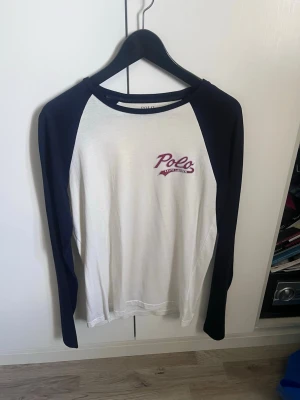 Långärmad t-shirt från Polo Ralph Lauren - Snygg långärmad t-shirt från Polo Ralph Lauren med vita paneler och mörkblå ärmar. Framsidan har ett broderat Polo-logotyp i vinrött och vitt. Klassisk basebollmodell med rund halsringning. Perfekt för en avslappnad och stilren look.