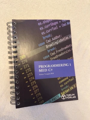 Programmering 1 med C# - Spiralbunden kursbok i programmering med C# av Krister Trangius, utgiven av Thelin Förlag. Omslaget är svart med kod i blått, vitt och gult. Perfekt för gymnasiekursen Programmering 1 eller för dig som vill lära dig grunderna i C#.