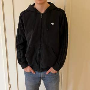 En svart Adidas hoodie med dragkedja. Kvaliten är bra. Storleken är uppskattningsvis medium alternativ liten large.