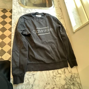 Ljussvart Calvin Klein tröja - En modern ljussvart Calvin Klein tröja med tryck. Tröjan är stretchig och mjuk som gör att man enkelt kan röra sig i den. Tröjan är av 100% bomull och har använts ett par gånger under året. Tröjan bibehåller dock fortfarande sin färg och passform. 