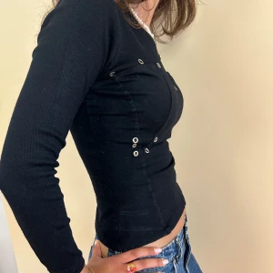 Långärmad topp med nitar - Långärmad topp med nitar som detaljer. Toppen formas efter kroppen och har korsettstruktur. Dragkedja på bakom 