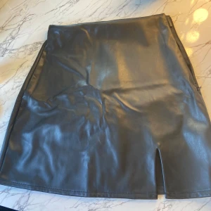 Svart kjol i skinn - En svart kortkjol i skinn med en liten slits framtill. Kjolen har en slät yta och en enkel design.