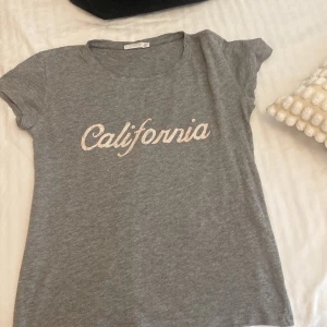 Grå t-shirt från Mango - En grå t-shirt från Mango med texten 'California' tryckt i vitt på framsidan. T-shirten har korta ärmar och en rund halsringning.💘