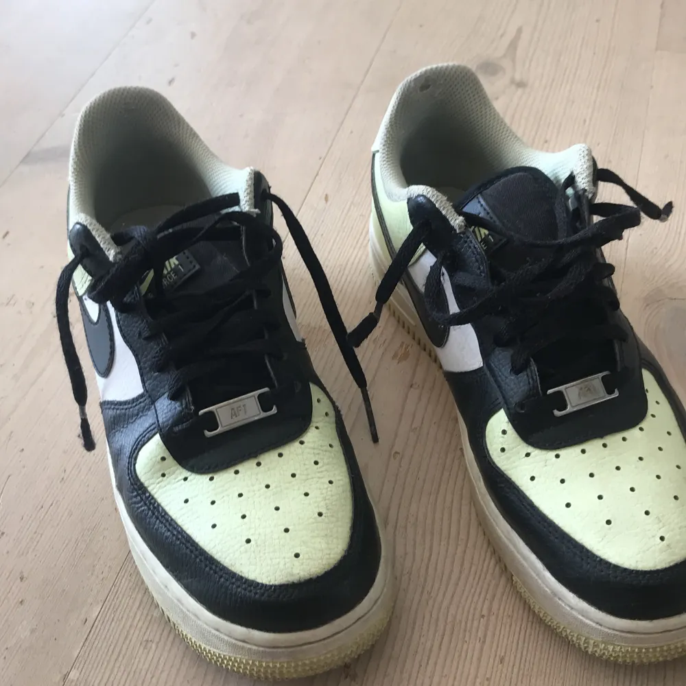 Ett par Nike Air Force 1 sneakers i svart, vit  och ljusgul färg. Skorna har snörning och en perforerad tåbox för ventilation. De är i storlek EUR 40, 25.5 cm. Skor.