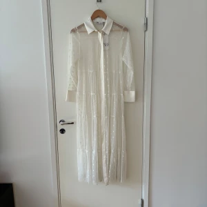 Vit långklänning från Zara - En elegant vit långklänning från Zara med prickigt mönster. Klänningen har långa ärmar och en klassisk krage. Den är genomskinlig och har knappar framtill.