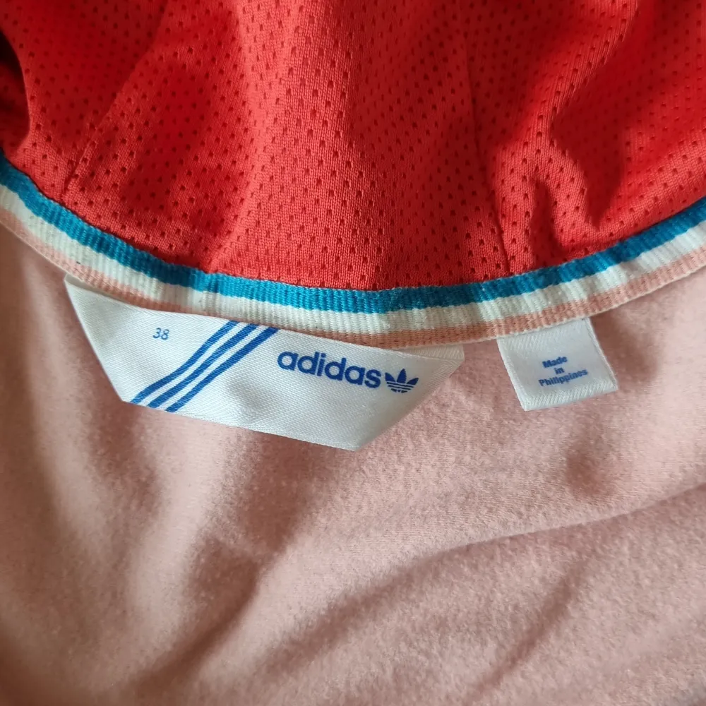 En färgglad jacka från Adidas med huva. Jackan har en kombination av blått, rosa och rött med dragkedja framtill. Den är långärmad och har ribbade muddar vid ärmslut och nederkant.. Hoodies.