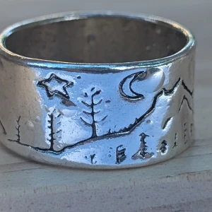 Silverring med naturmotiv - En bred silverring med ingraverade naturmotiv som inkluderar stjärnor, måne, träd och berg. Ringen har en rustik och handgjord känsla.ny