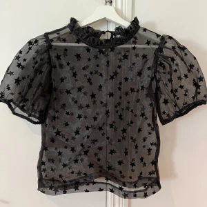 Svart transparent blus med stjärnmönster - Säljer en superfin svart transparent blus med stjärnmönster. Blusen har korta puffärmar och en söt volangkrage. Perfekt för festliga tillfällen eller när du vill känna dig extra speciell. Passar bra med ett linne under eller som den är för en mer vågad look. 🖤