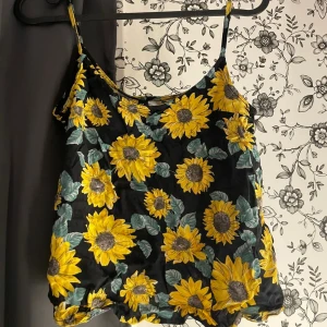 Svart linne med solrosmönster - Säljer ett svart linne med ett färgglatt solrosmönster. Linnet har smala axelband och är perfekt för sommaren. Materialet känns lätt och luftigt, vilket gör det idealiskt för varma dagar. Passar bra till både jeans och shorts!