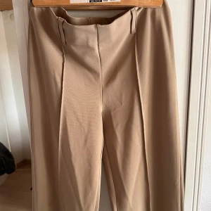 Beige kostymbyxor - Säljer ett par snygga beige kostymbyxor. De är högmidjade och har en rak passform. Perfekta för både jobb och fest! Byxorna är i nyskick och har en dragkedja framtill. Materialet känns som bomull eller en bomullsblandning, vilket gör dem bekväma att bära hela dagen.