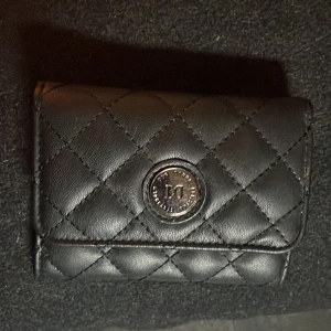 Svart quiltad plånbok från Don Donna - Säljer en snygg svart plånbok från Don Donna. Den är quiltad och har en liten metallogo på framsidan. Plånboken har flera fack för kort och en dragkedja på baksidan för mynt. Den är i bra skick och perfekt för att hålla ordning på dina pengar och kort. Köp gärna med köp nu!💕