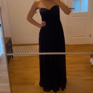 Mörkblå axelbandslös klänning - Mörkblå axelbandslös klänning med en hjärtformad urringning. fint fall och lång, går över fötterna på mig som är 178cm.  Har ett  silikonband på insidan fram så den sitter uppe ganska bra. Stängs med dold dragkedja i sidan. Använd en gång men ser ut som ny. 