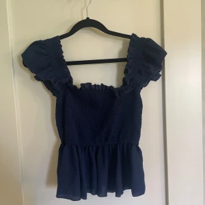 Mörkblå topp med volangärmar - Säljer en söt mörkblå topp med volangärmar. Toppen har en smockad överdel som ger en tight passform och en volangkant nedtill som ger en fin detalj. Perfekt för sommaren och festliga tillfällen!